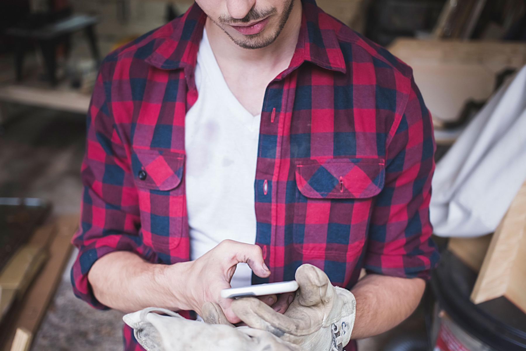 A tradesperson in a work shirt, gloves in one hand, checking a phone in a workshop.